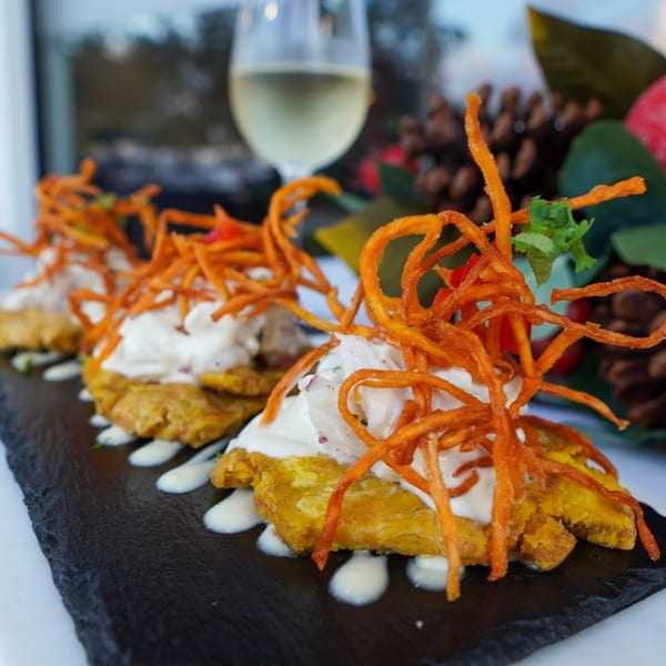 Peruvian tostones topped with white sauce and crispy orange strips, served on a black slate.