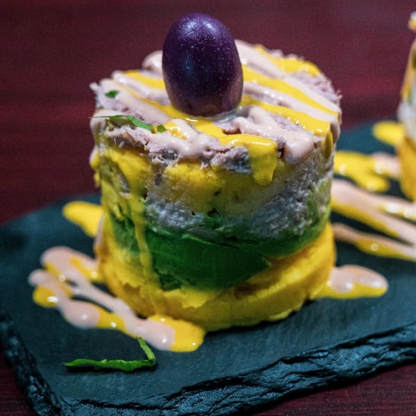 Peruvian causa dish with layered yellow potato, green avocado, and tuna, topped with sauces and an olive.