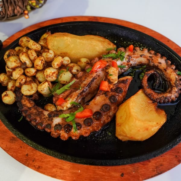 Grilled octopus with roasted potatoes and corn, garnished with red peppers and herbs, served on a hot plate.